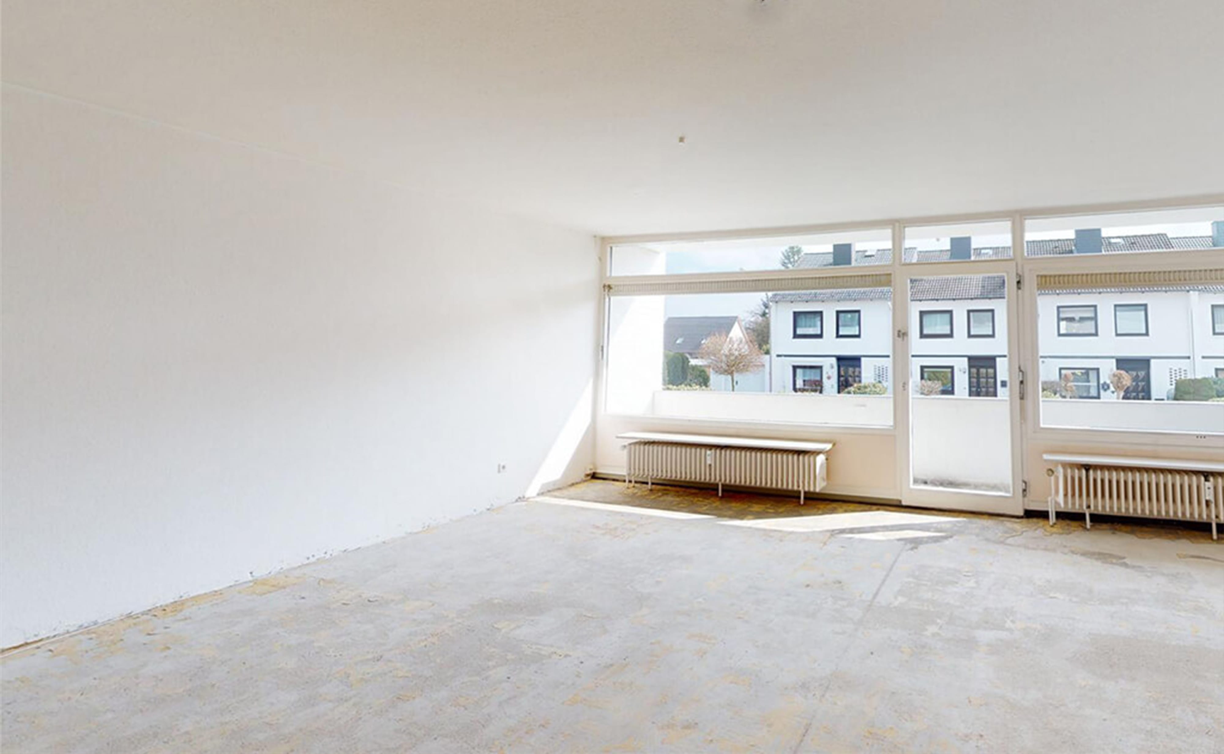 Vorher-Bild für virtuelles Home Staging: Ein leerer, renovierungsbedürftiger Raum mit rohem Estrichboden, weißen Wänden und einer großen Fensterfront mit alten Heizkörpern vor der Sanierung.